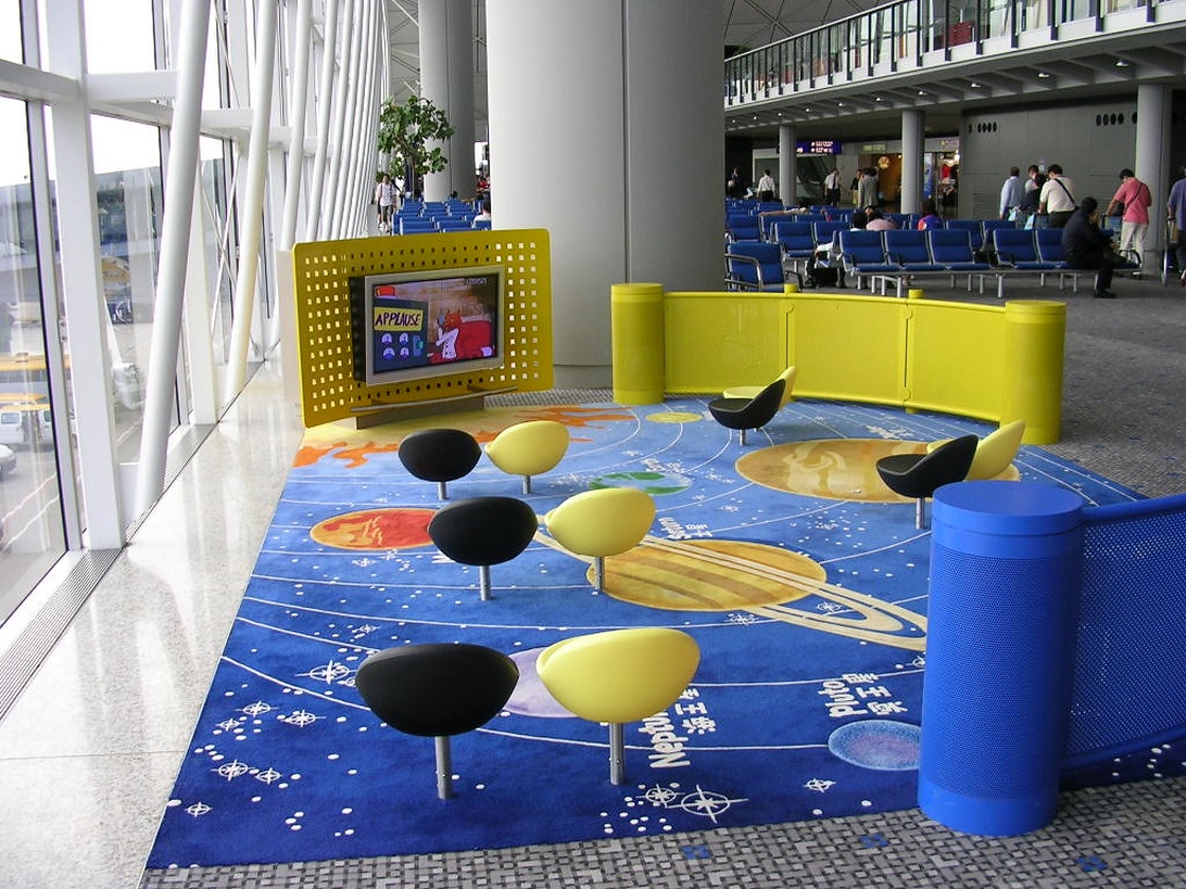 Een kleurrijke speelruimte voor kinderen met een blauw tapijt met ruimtethema, gele en zwarte capsule-achtige stoelen en een geel mediatiostation.